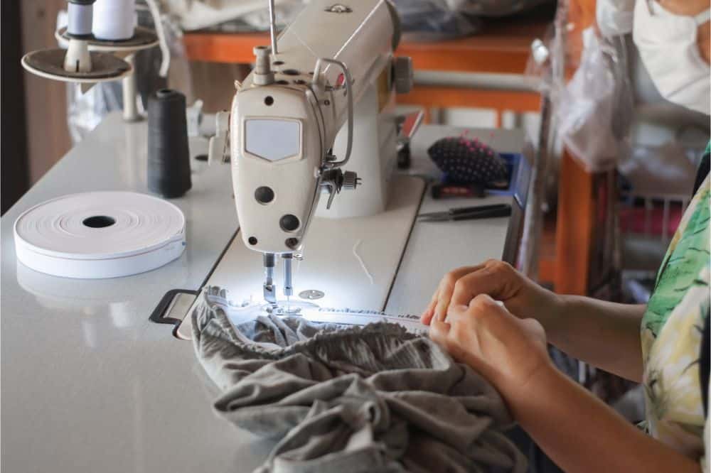 Hand of seamstress is using the white industrial sewing machine to sew elastic strips of pants close-up