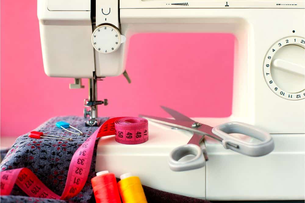 Fabric, measuring tape, thread, scissors and sewing machine on a pink background