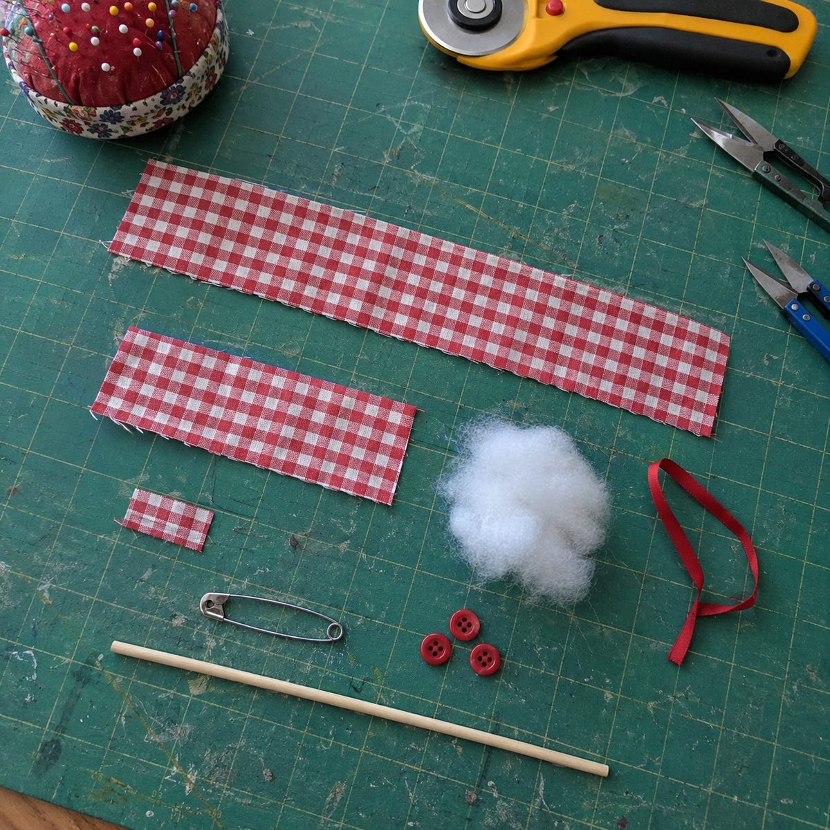Cut gingham strips, fiberfill, ribbon, safety pin and buttons for a mini fabric wreath laid out on a cutting mat