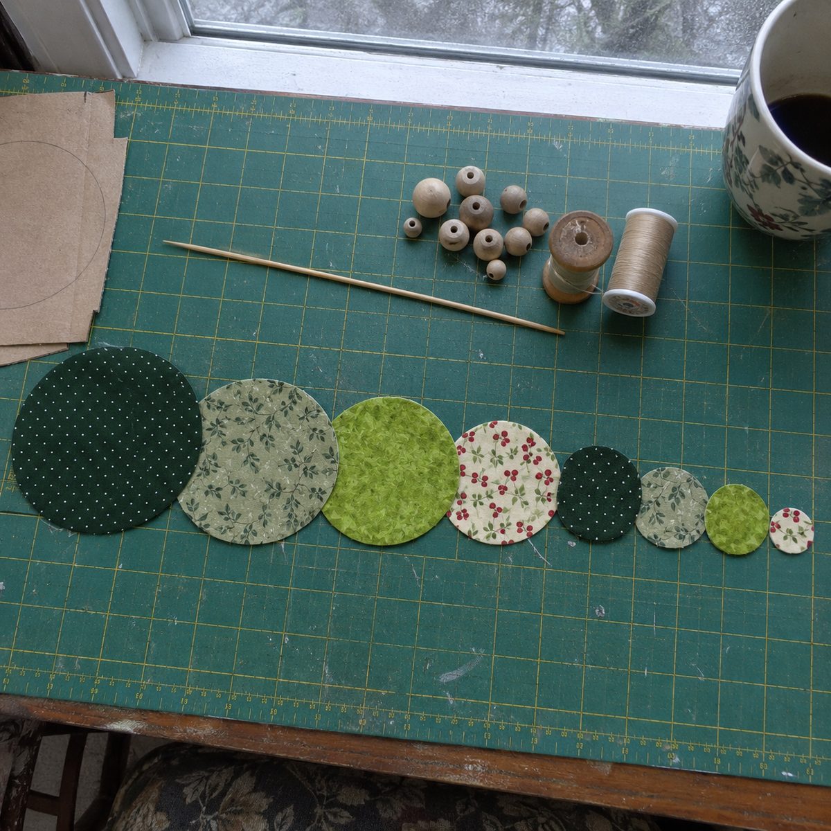 Nine green fabric circles cut in graduated sizes laid out on a cutting mat with a skewer, wooden beads and a thread spool