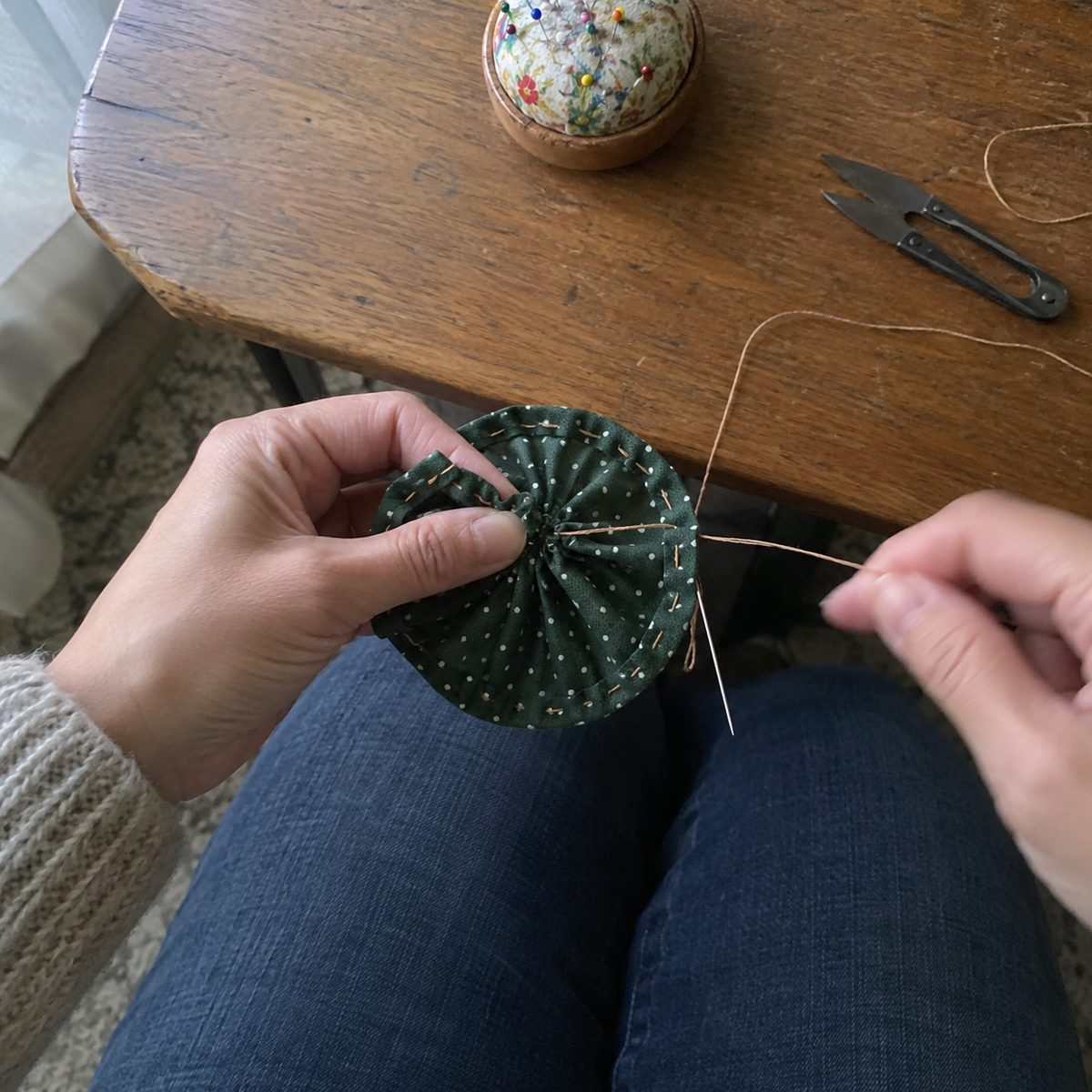 Hands pulling the gathering thread on a green fabric circle to form a yo-yo, with the folded hem and running stitches showing