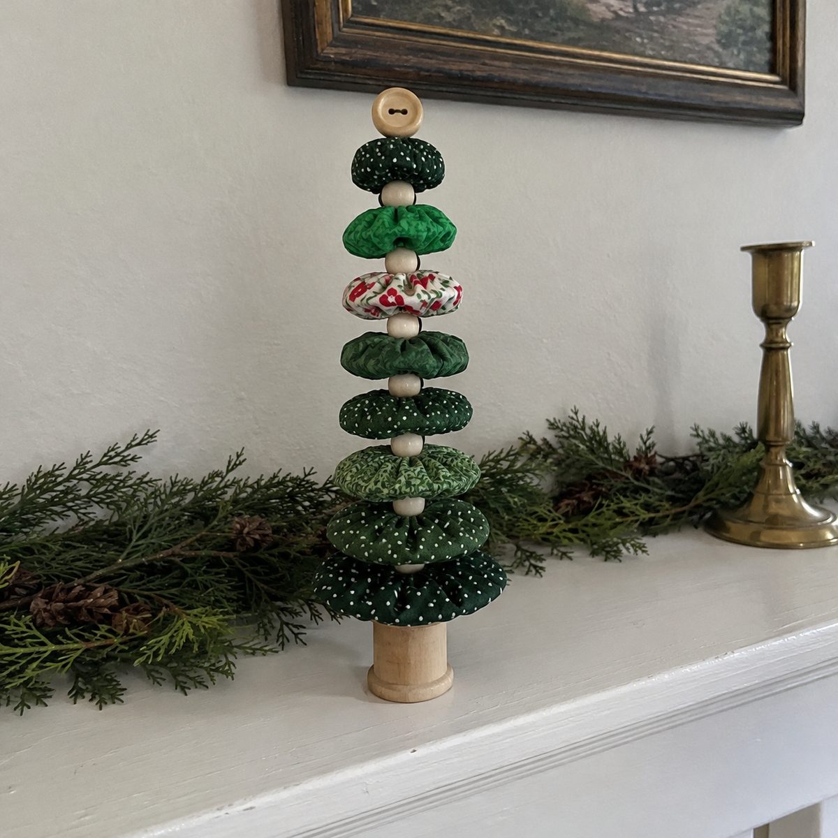 Finished fabric yo-yo Christmas tree in mixed green prints with a button top standing on a wooden spool on a mantel