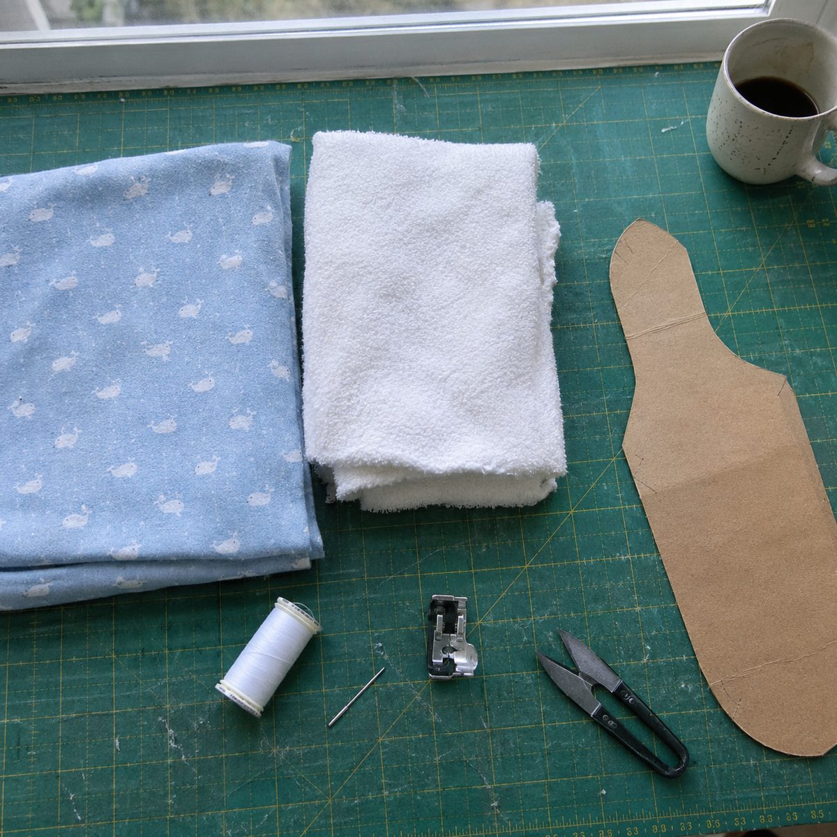 Burp cloth materials on a cutting mat: blue whale flannel, white terry cloth, thread, walking foot and a cardboard contoured template