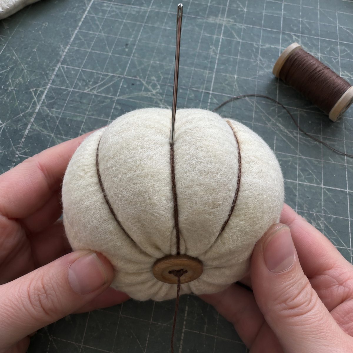 Close-up of a doll needle pulling segments through a fabric pumpkin with a button anchor on the bottom