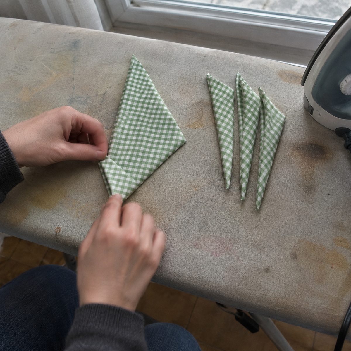 Hands folding the outer edge of a gingham cotton square into a narrow point on an ironing board