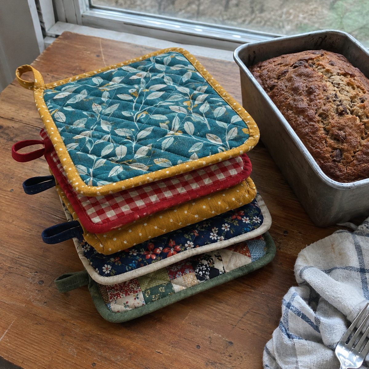 A stack of finished handmade quilted pot holders in assorted fabrics on a kitchen table